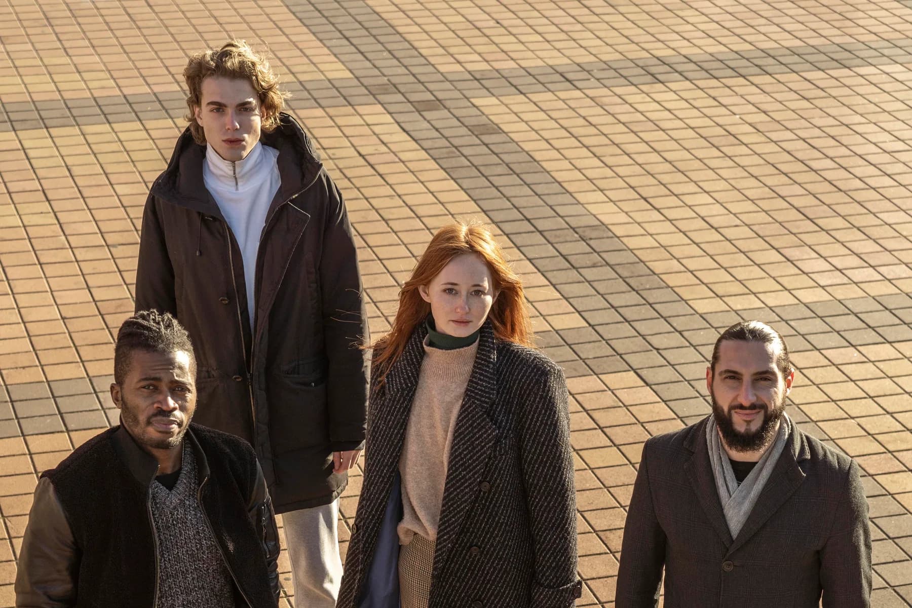Four cast members from above, crossing a tiled square in low sun.