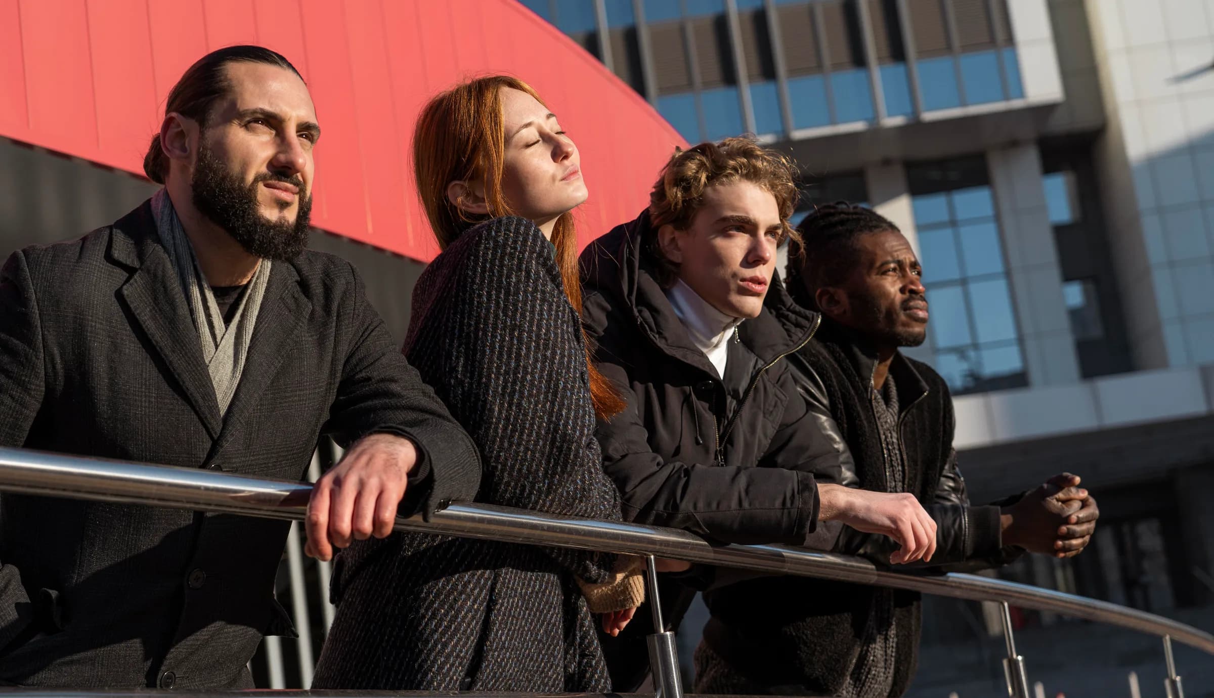 Four cast members at a rail in low sun, looking past the camera.